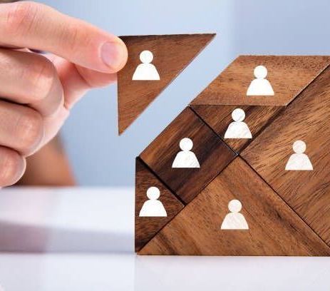 Hand placing a person-shaped block into a wooden puzzle depicting a team, symbolizing recruitment or collaboration.