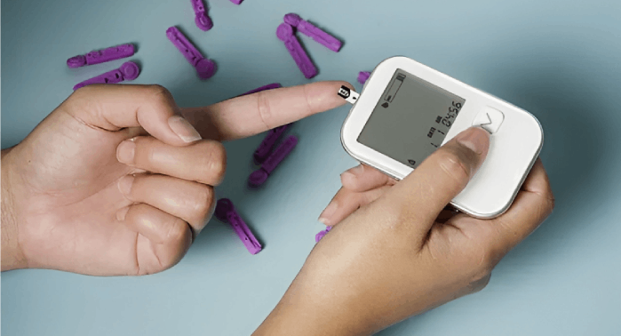 Person using a blood glucose meter to test their finger. Purple lancets scattered on blue surface.