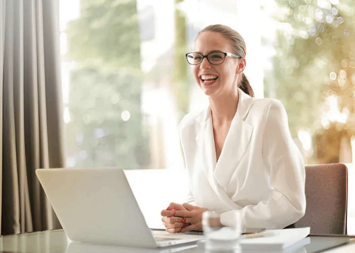 Woman in white suit, glasses, and ponytail laughs, sitting at a desk with a laptop and glass in front of a window.