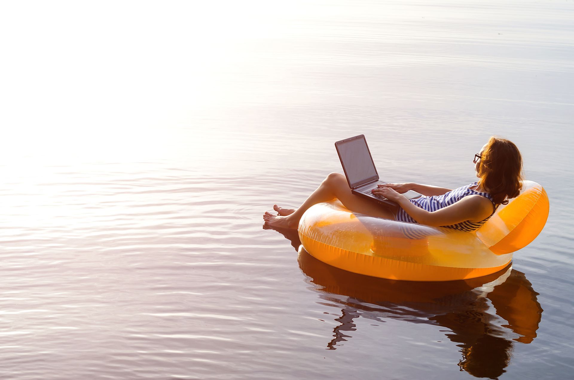 Woman on an inflatable ring in water working on laptop, sunny day.