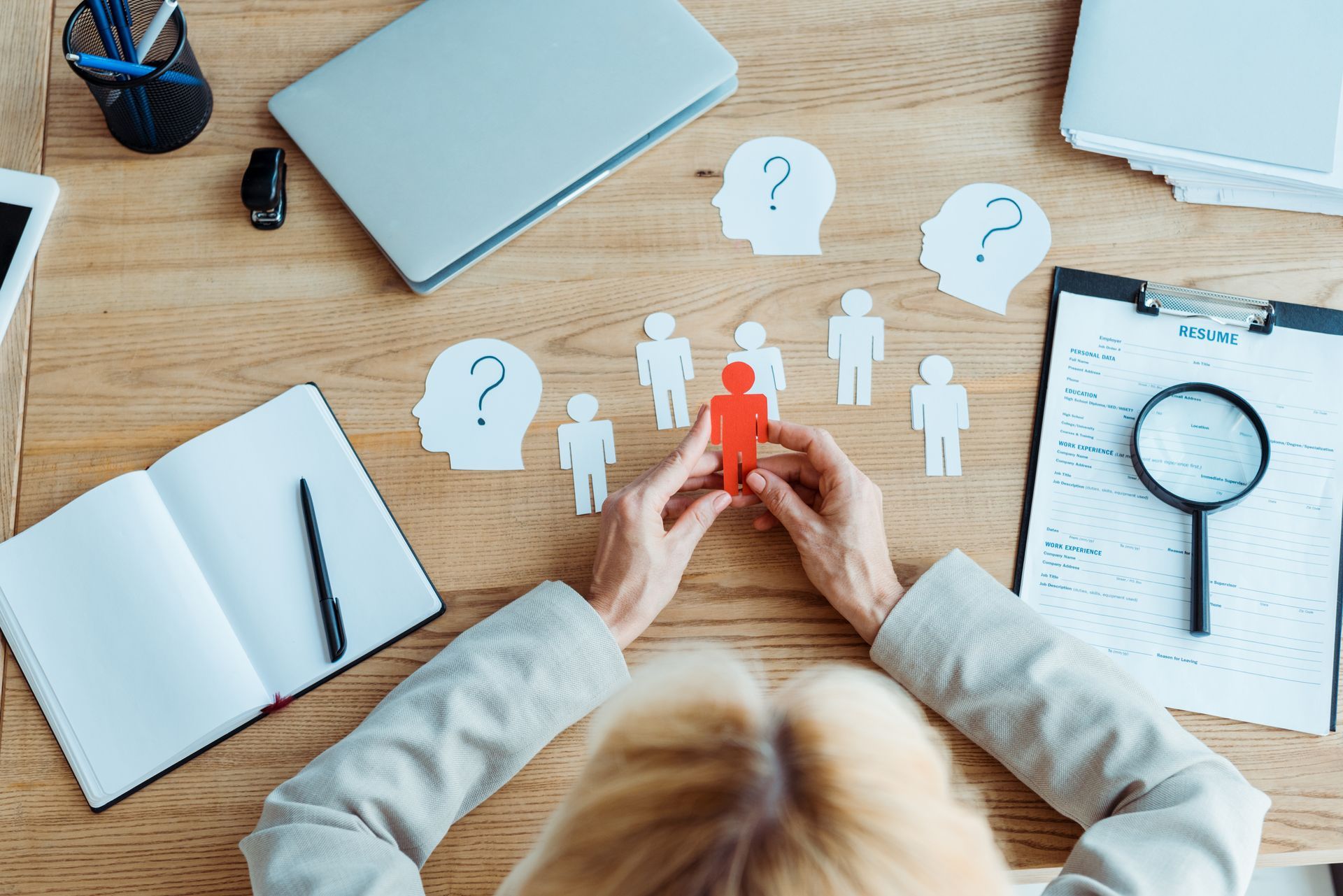 Hands holding red figure among other figures and question marks on desk with laptop, notepad, and magnifying glass