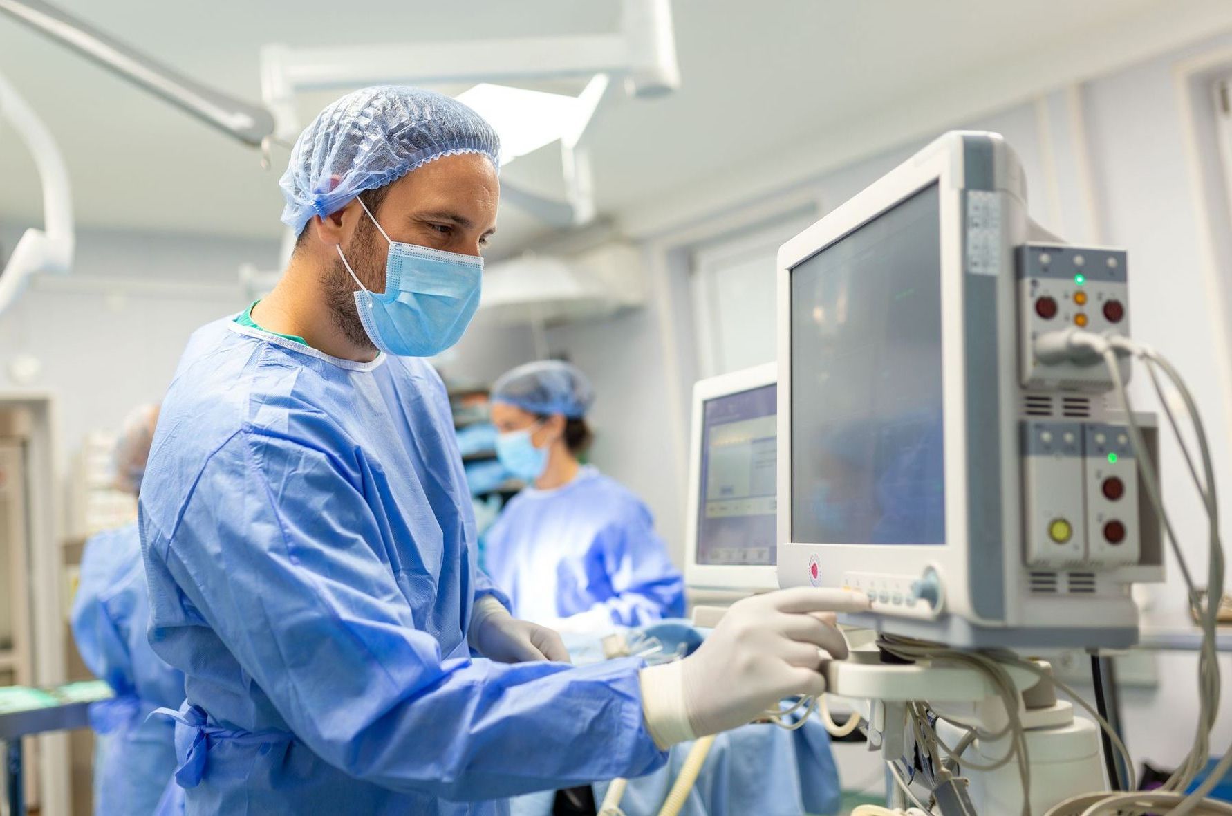 Surgeon in blue scrubs, mask, and gloves adjusts medical monitor in operating room; another medical worker in background.