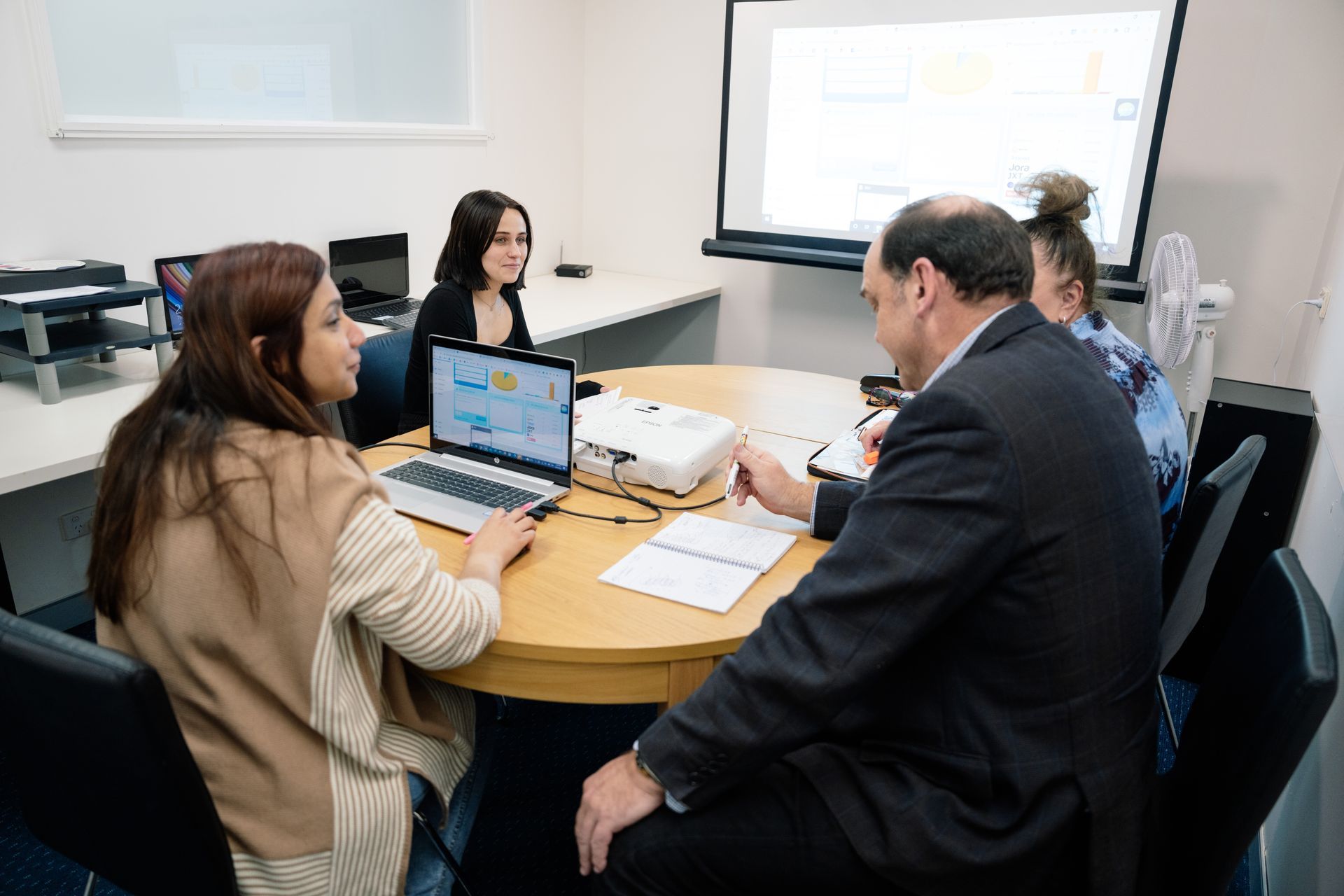 Four people around a table in a meeting room, reviewing papers and a laptop, projector in center.
