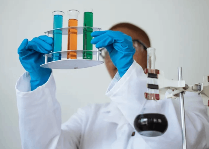 Scientist in lab coat and gloves holding a rack of colorful test tubes.