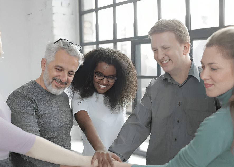 Diverse group of people with hands together, smiling and united, in a bright indoor setting.