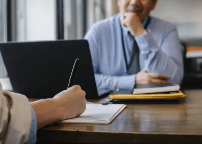 Person writing on paper at a desk, interviewing another person, laptop, and files in the background.