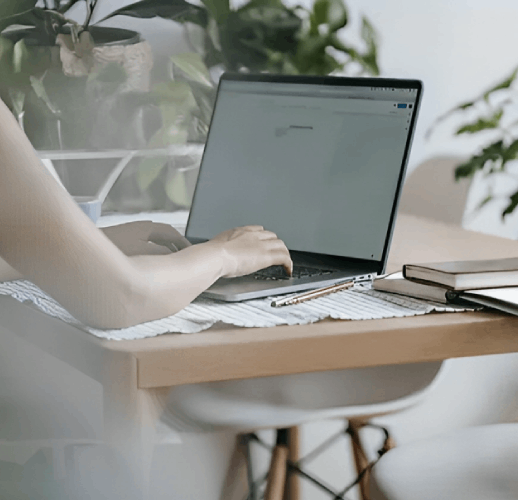 Person typing on a laptop at a desk; books and a pen are nearby.
