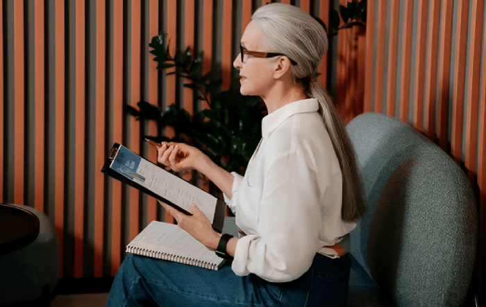 Woman with gray hair in white shirt and jeans reviews a document in a waiting area.