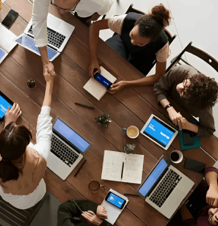 People at a wooden table, laptops, tablets, and phones. Two shake hands, others work, coffee present.