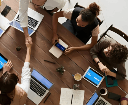 People in a meeting around a wooden table with laptops, tablets, and a handshake.