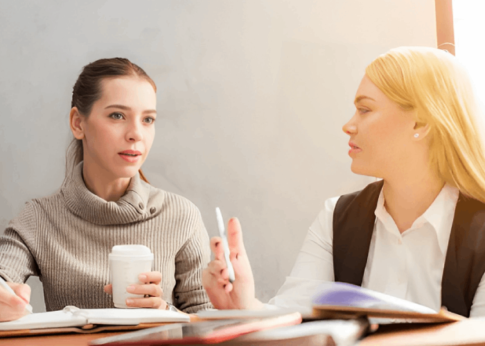Two women at a table, one holding a coffee cup, the other with a pen, engaged in a conversation.