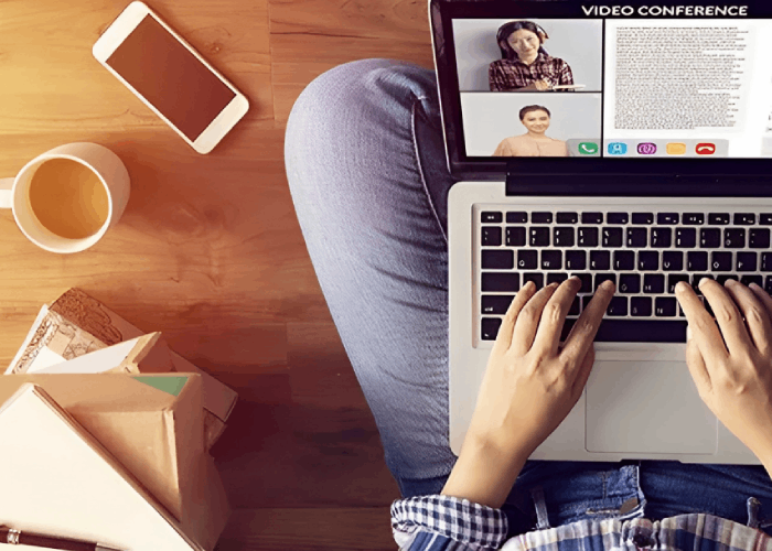 Person on laptop in video conference, working from home, with coffee and phone nearby.