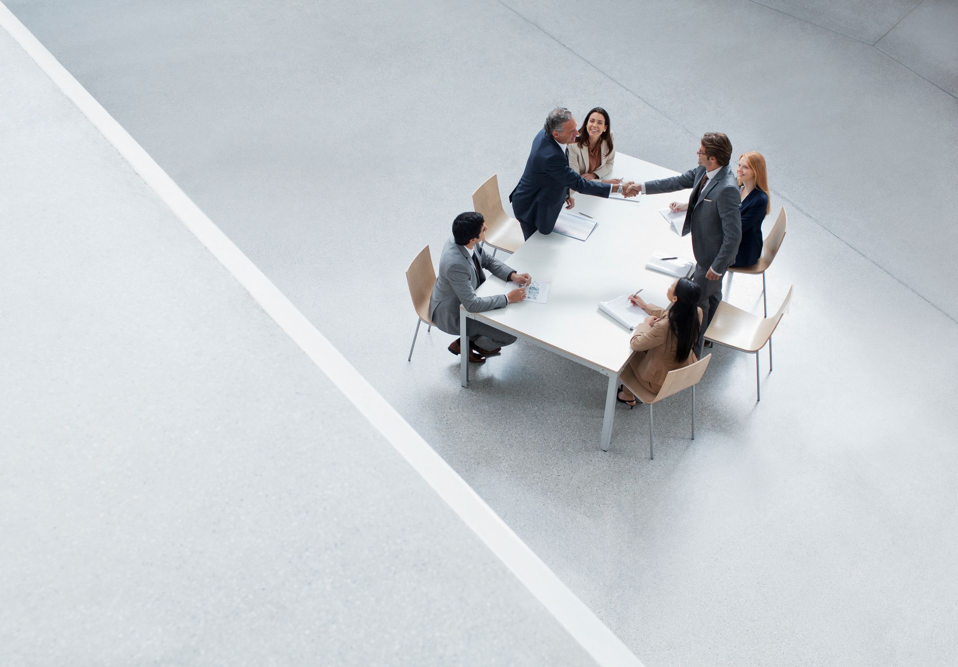 Businesspeople in suits at a white table shaking hands, in a modern, bright office setting.