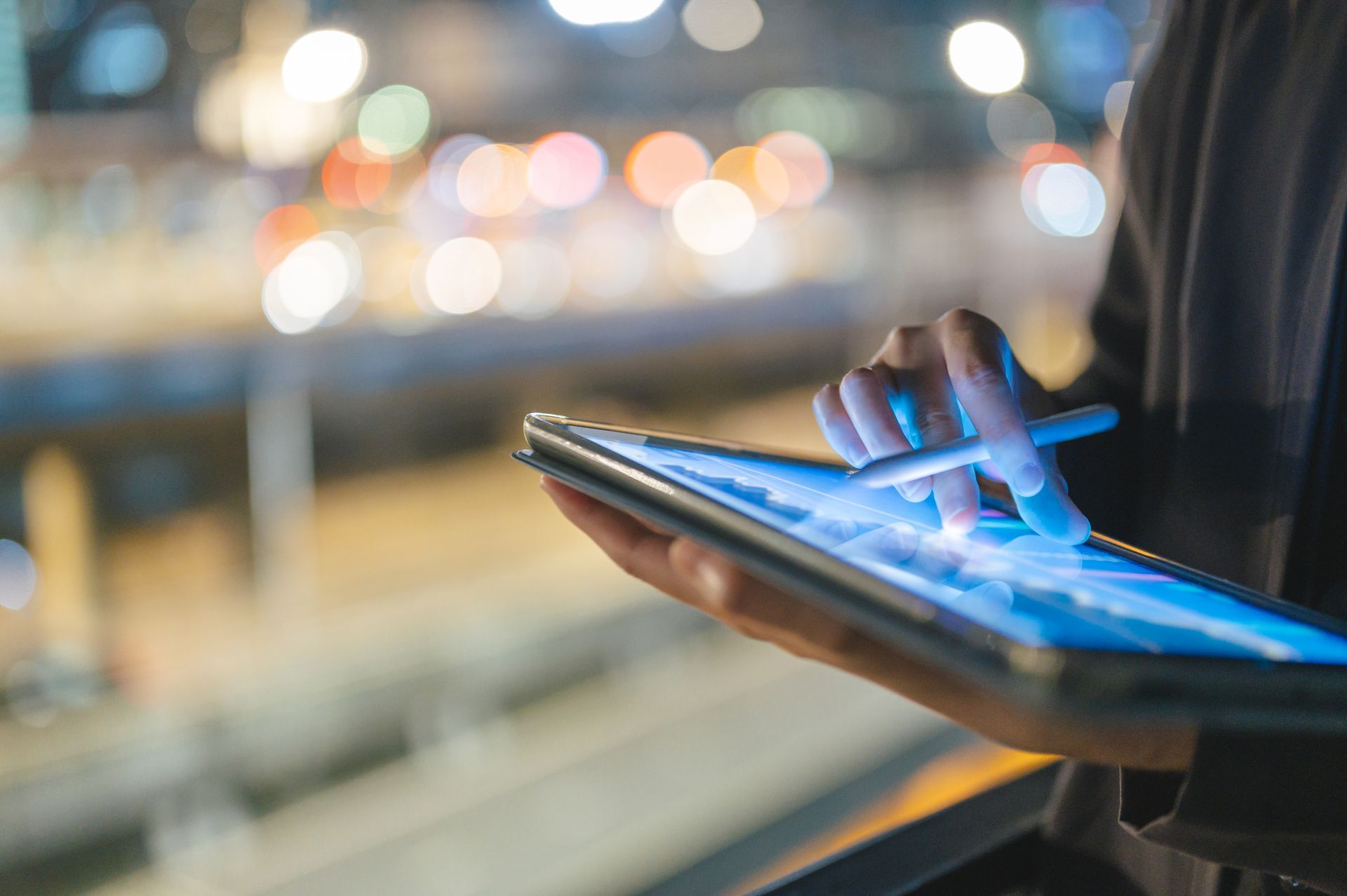 Person using a stylus on a glowing tablet screen at night, city lights blurred in background.
