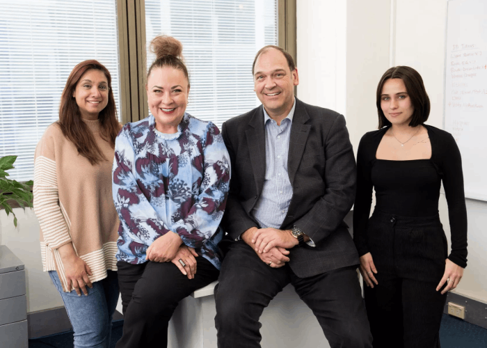 Four people posing for a photo in an office setting; smiling, one person seated, neutral background.