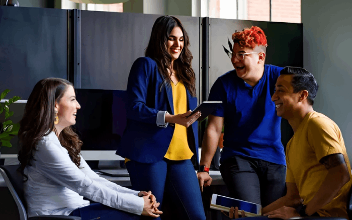 Four diverse people in an office discussing a tablet, laughing. Woman in blue blazer.