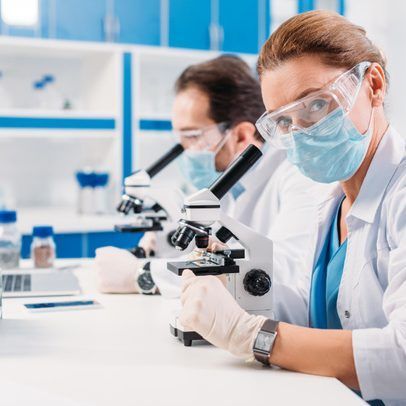 Two scientists wearing masks, goggles, and lab coats look at microscopes in a lab.