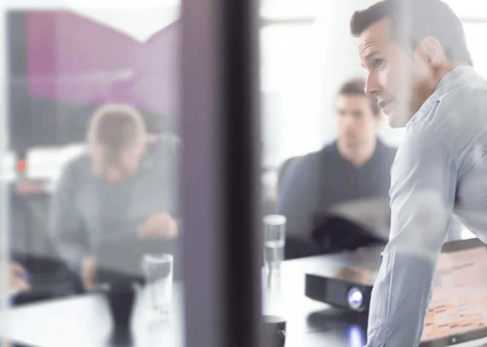 Man in a meeting, leaning forward, looking intently. Others blurred in background, office setting.