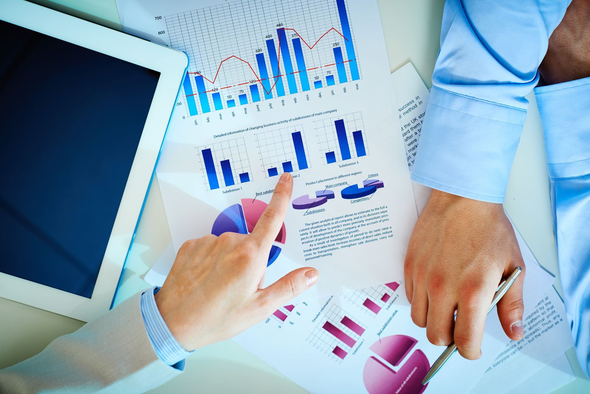 Hands pointing at charts and graphs on a table, analyzing data. Blue and pink data visualized.
