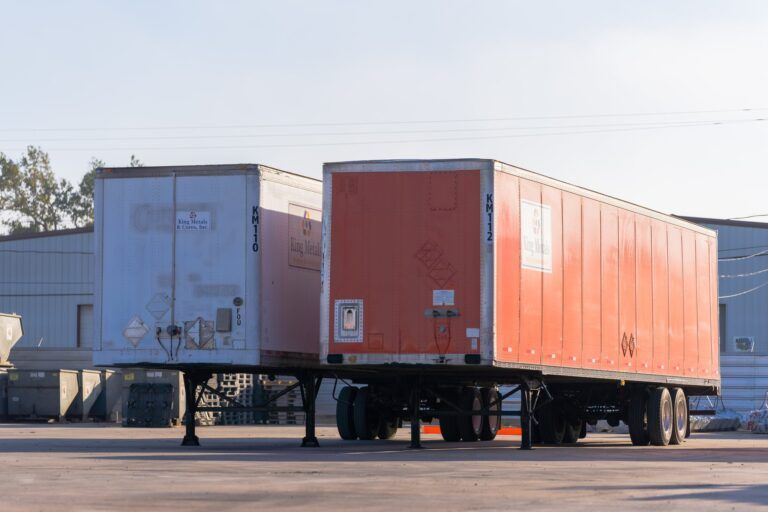 Two semi-trailers parked side by side on a lot, one white and one orange, viewed from the rear.