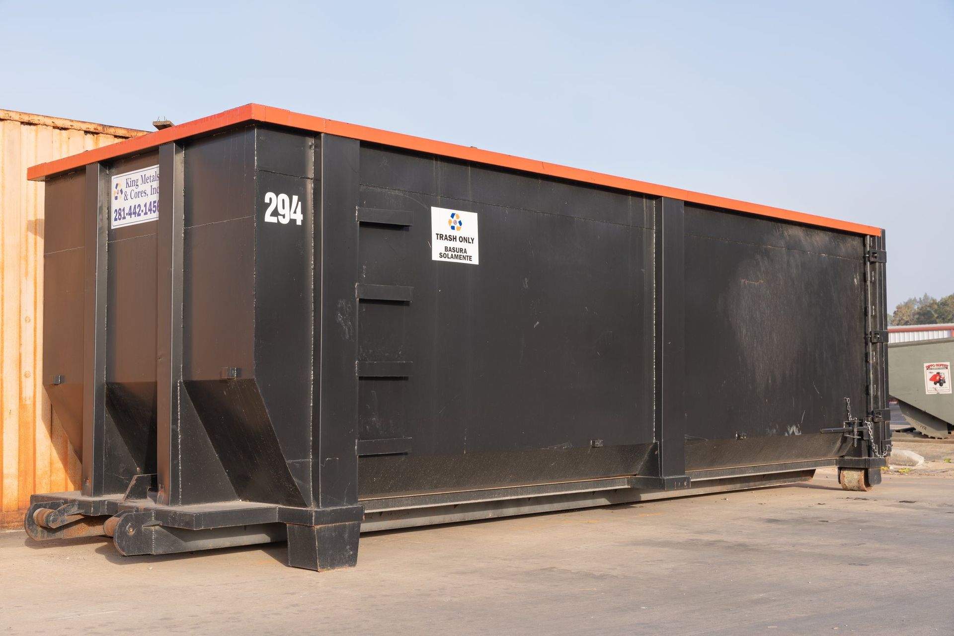 Large black dumpster with orange lid in an outdoor industrial area