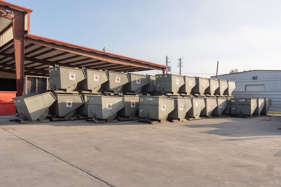 Stacked gray dumpsters beside a brick building on a sunny concrete lot
