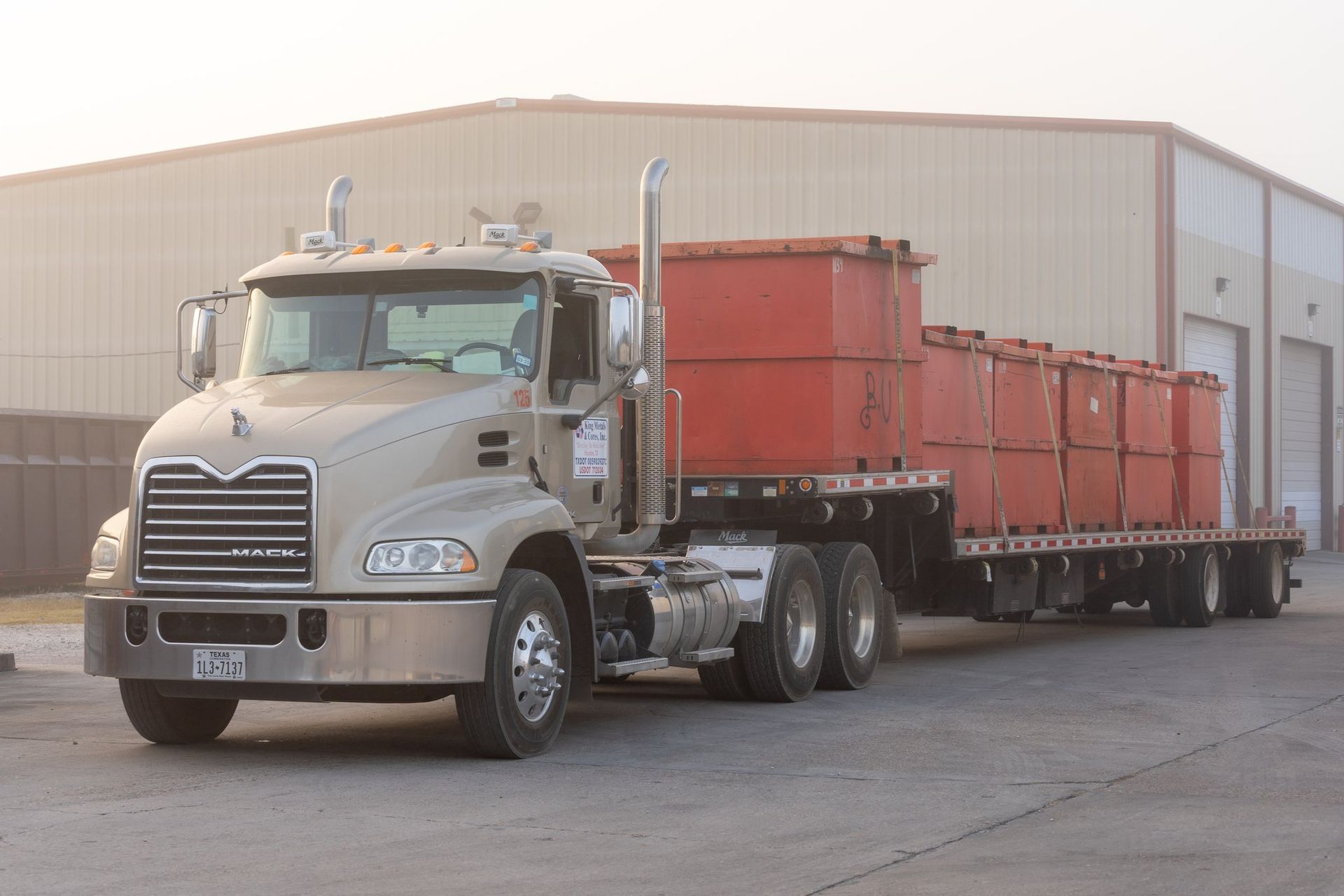 Silver semi-truck hauling red trailers parked outside a large warehouse