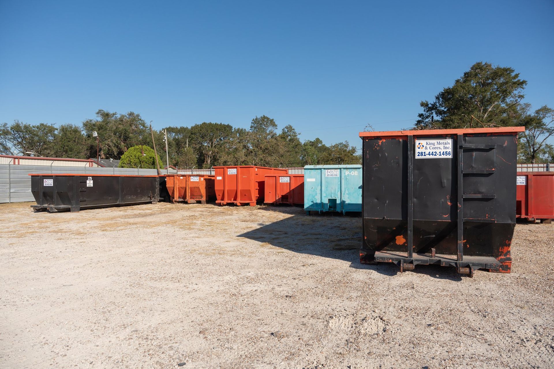 Row of orange and black dumpsters in a gravel lot under a clear blue sky