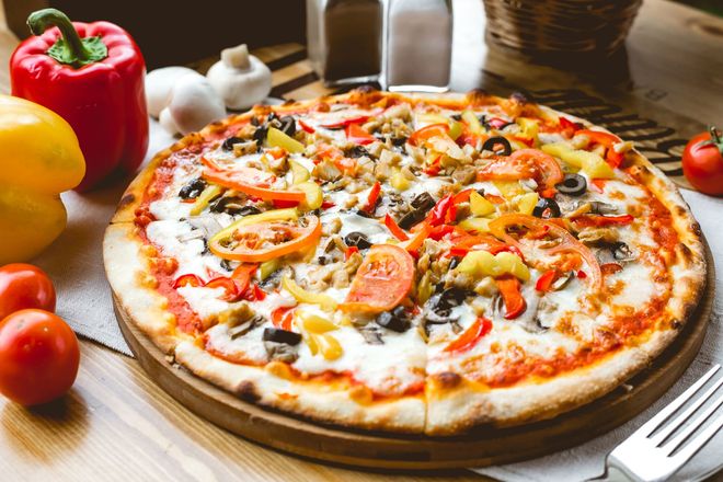 Cheese pizza topped with mushrooms, peppers, and tomatoes on a wooden board with fresh vegetables nearby