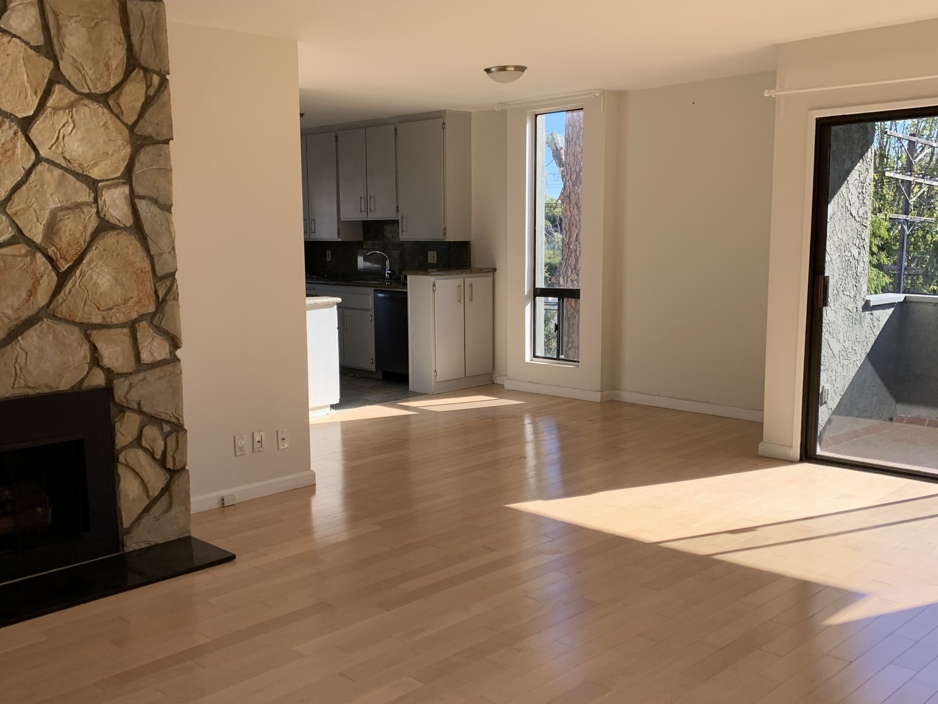 Empty living room with stone fireplace, hardwood floors, and bright sunlight from large windows.