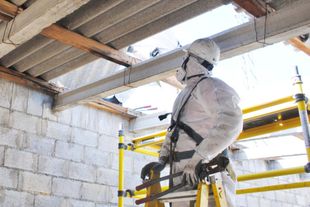 Worker in white protective suit inspecting a ceiling opening from a yellow scaffold inside a construction site
