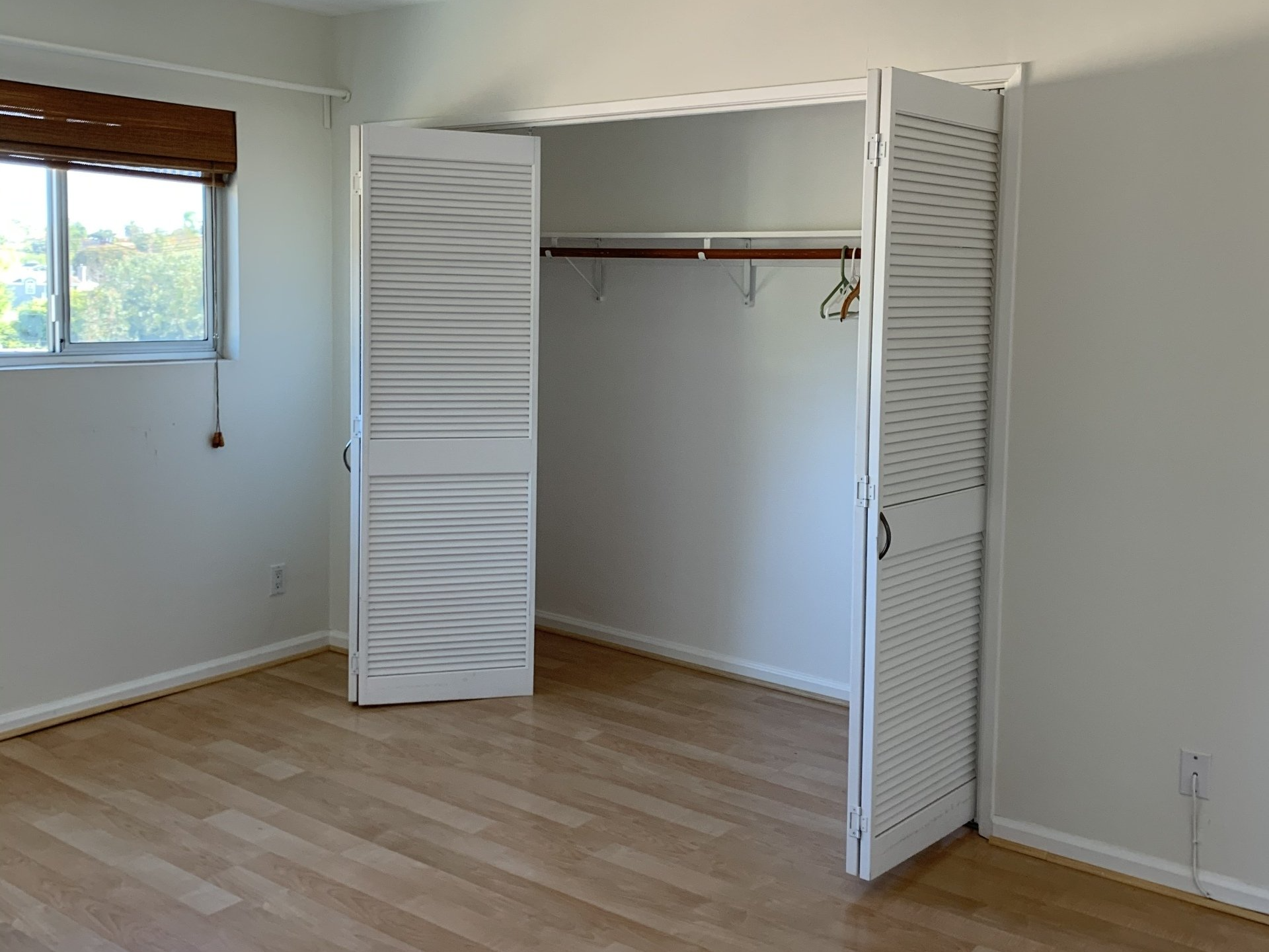 Empty bedroom with open white louvered closet doors, wood floors, and a window with blinds.