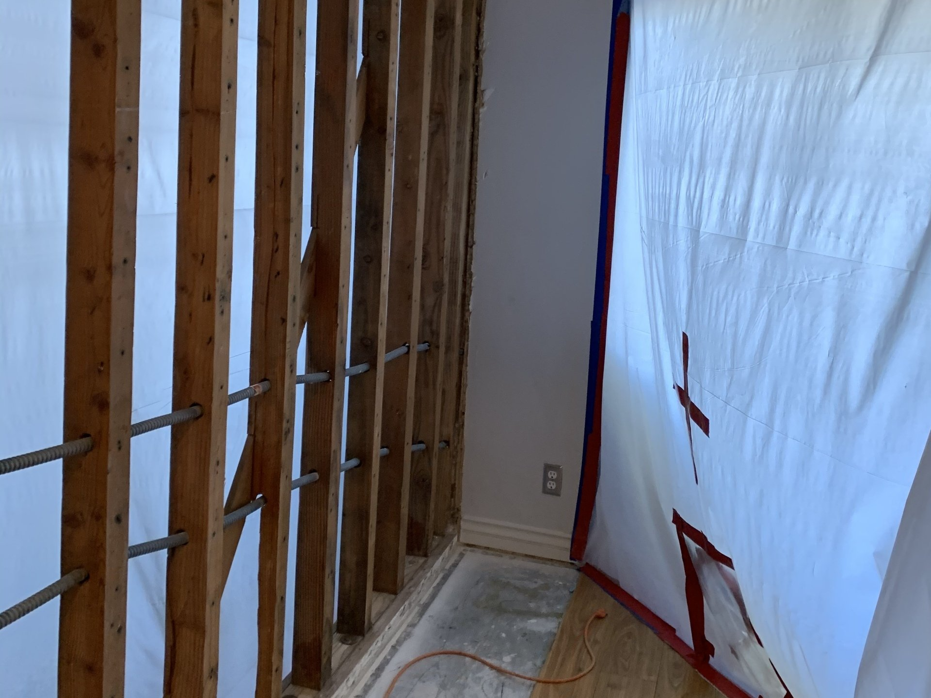 Exposed wall studs in a gutted room with white plastic sheeting and exposed flooring, under renovation.