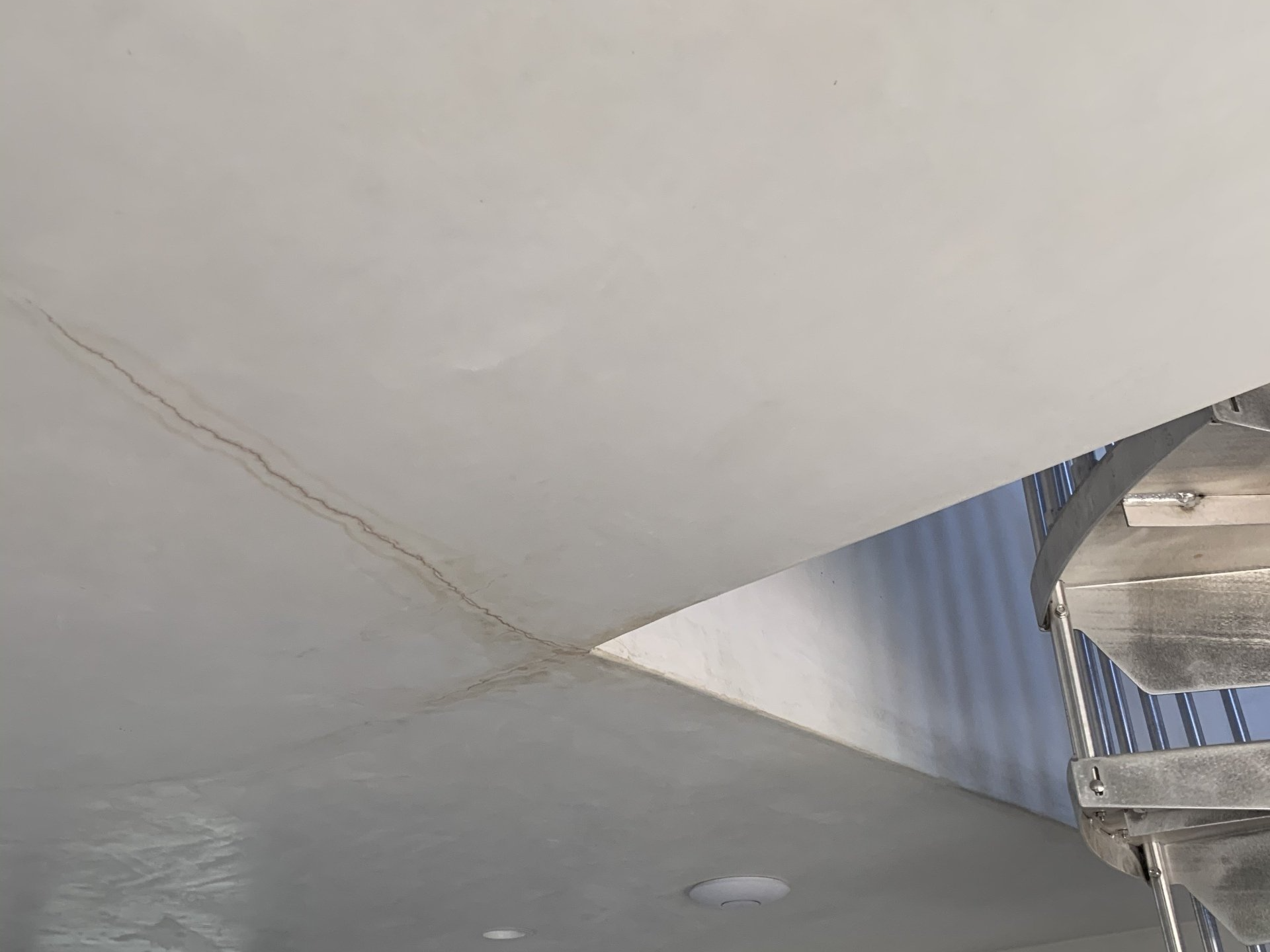 Close-up of a cracked white ceiling with a small gray wall vent and a metal fixture on the right.