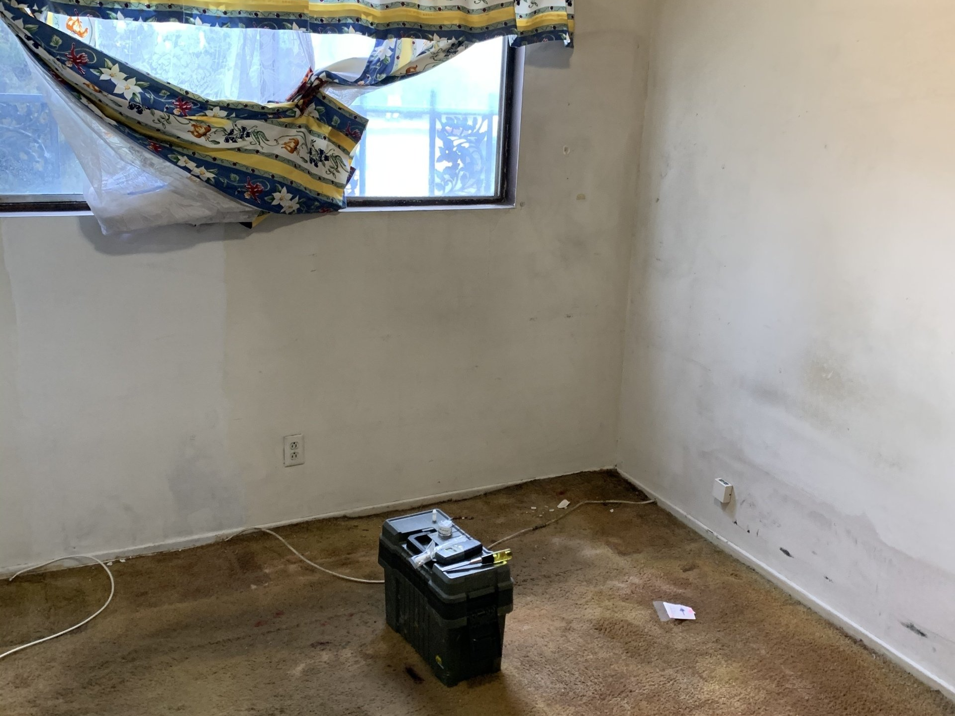 Empty room with a broken window covering, white walls, and a black toolbox on the floor.