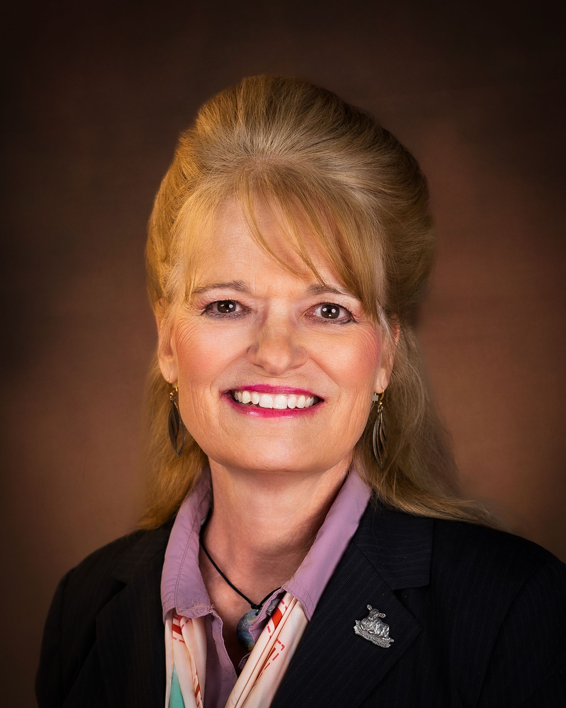 Woman with blonde hair smiles, wearing a suit and floral scarf, brown background.