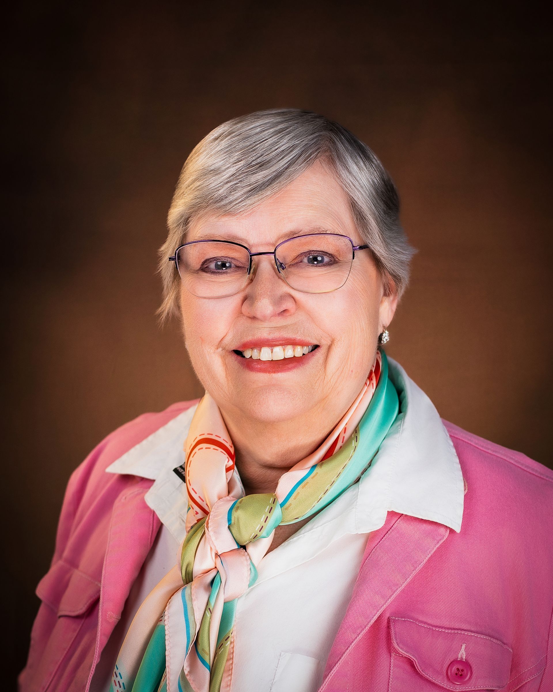 Woman with blonde hair smiling, wearing a dark blazer and green shirt, brown background.