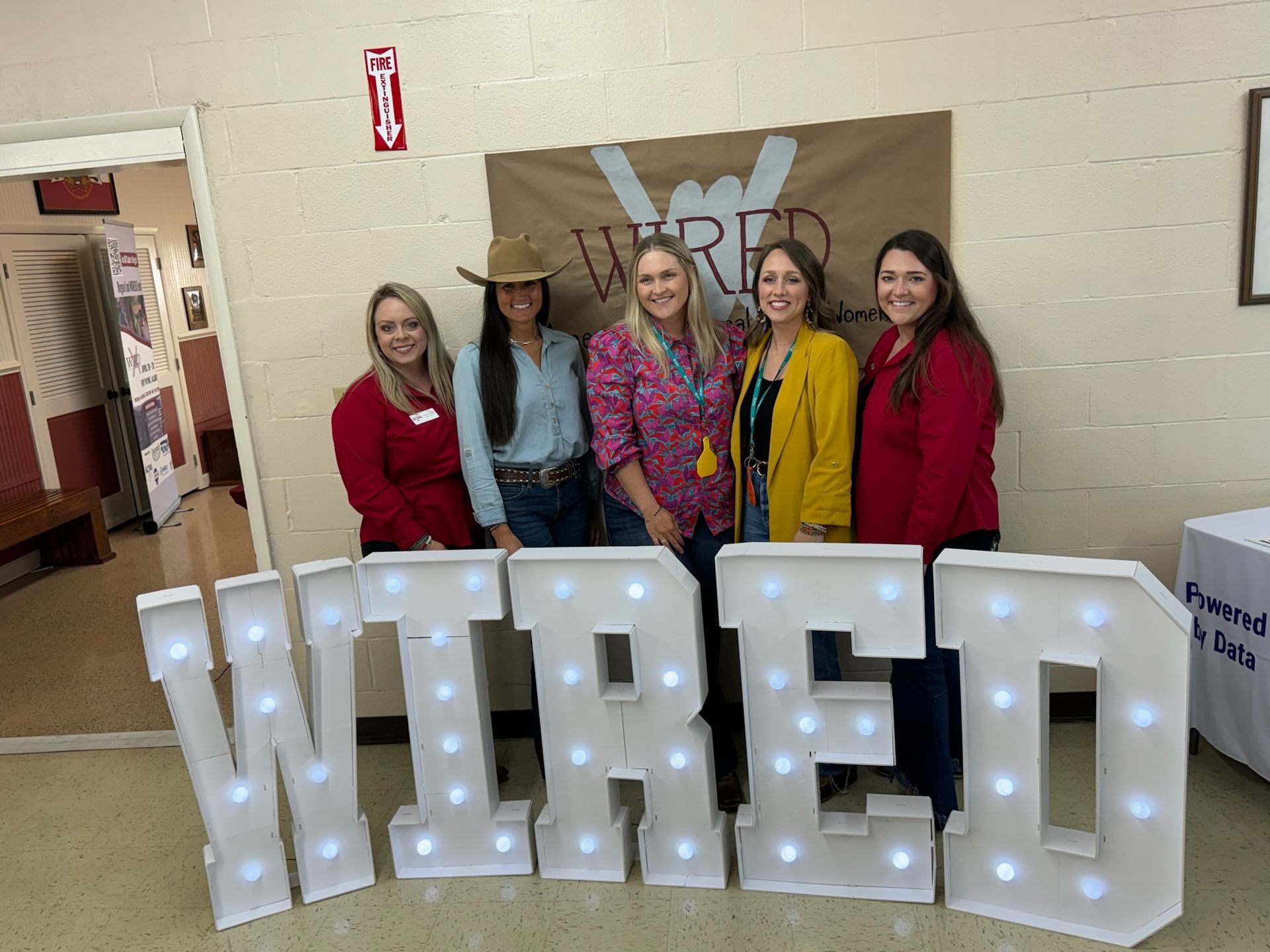 Five women smiling, posing behind illuminated 