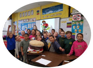 Students in a classroom with a giant burger, excited faces. Teacher with a cartoon on the whiteboard.
