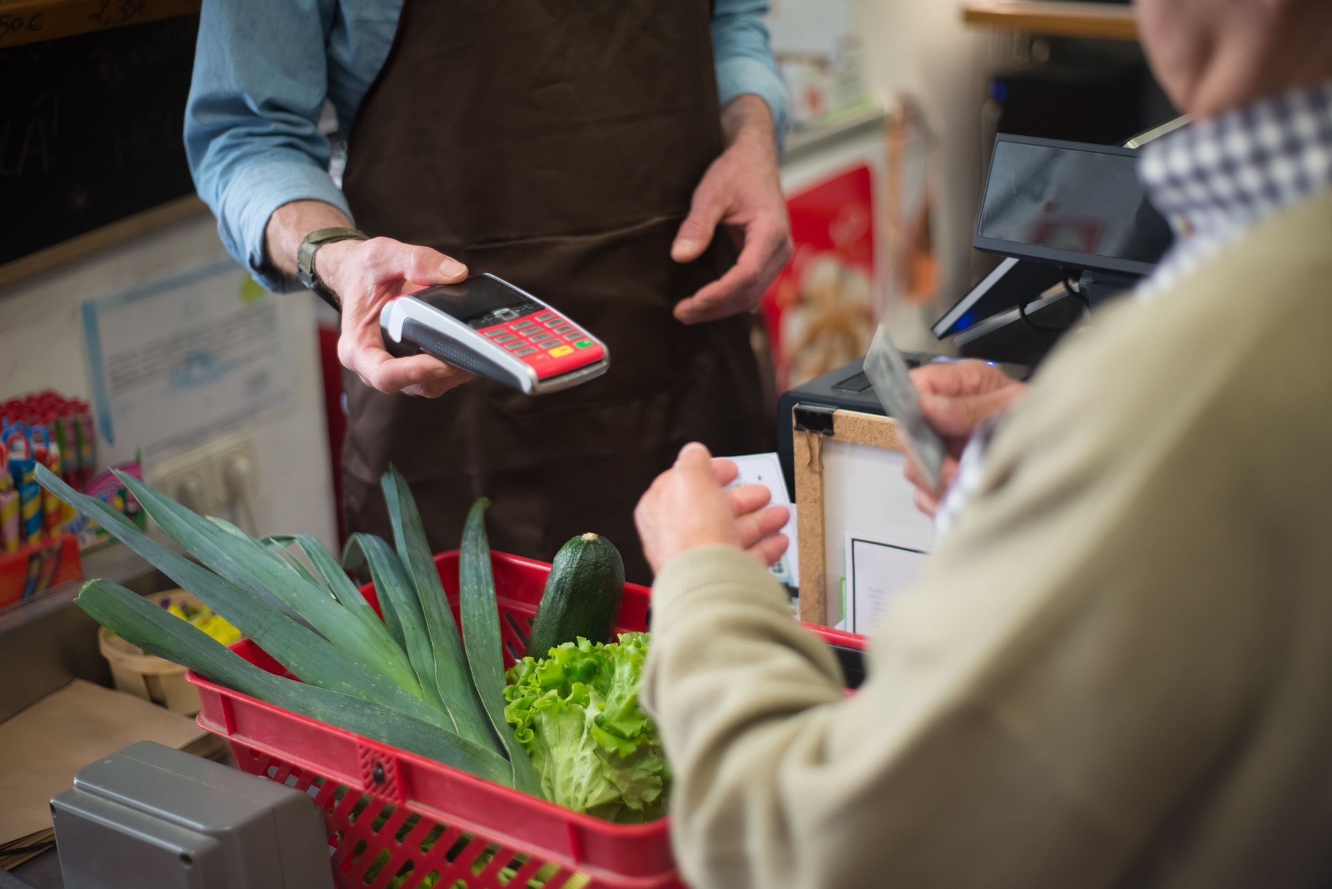 Customer paying by card at a grocery checkout with fresh vegetables in a basket