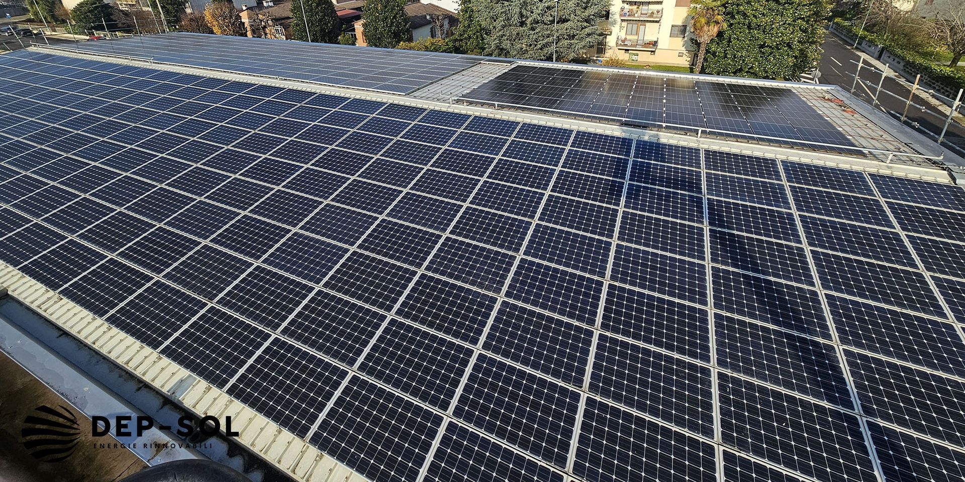 Pannelli solari che ricoprono un tetto in pieno sole, con case e alberi sullo sfondo.