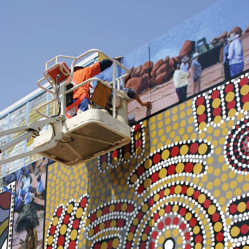 A Man in a Bucket is Painting a Mural on the Side of a Building — Bluey's Signs in Ciccone, NT A Man in a Bucket is Painting a Mural on the Side of a Building — Bluey's Signs in Ciccone, NT