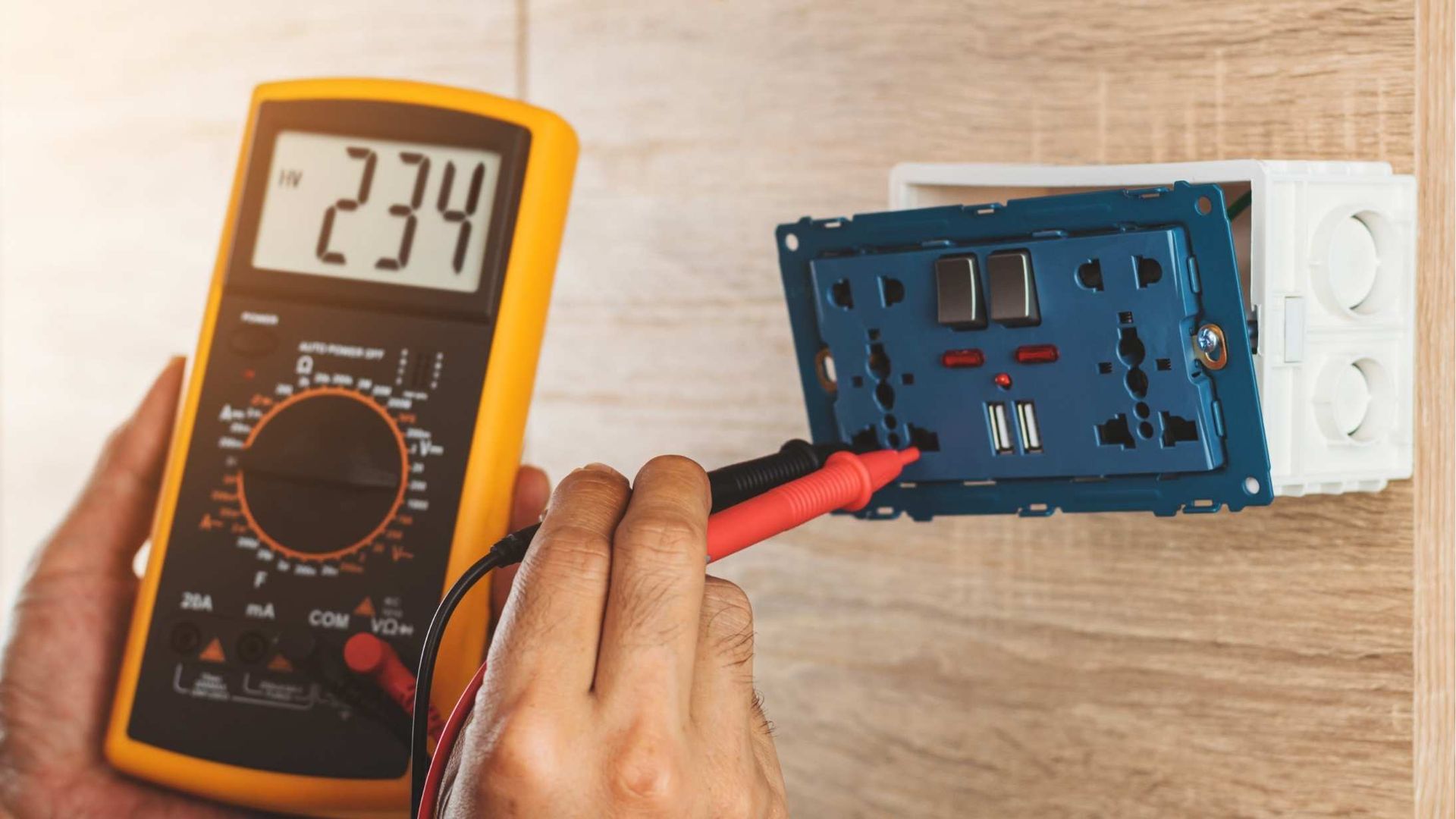 Handheld multimeter testing a blue electrical outlet mounted on a wooden wall