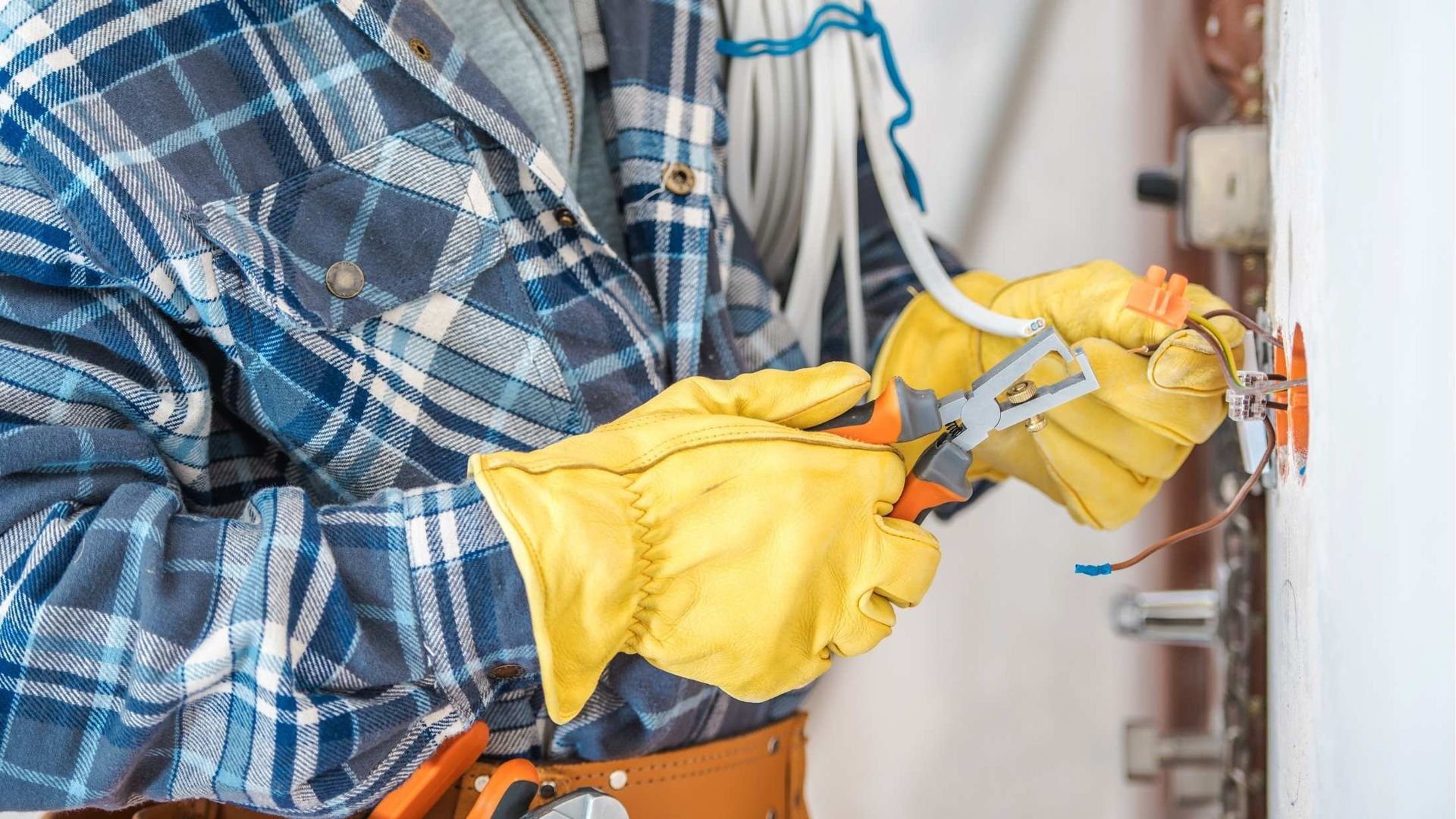 Electrician in blue plaid shirt and yellow gloves using pliers on wall wiring.