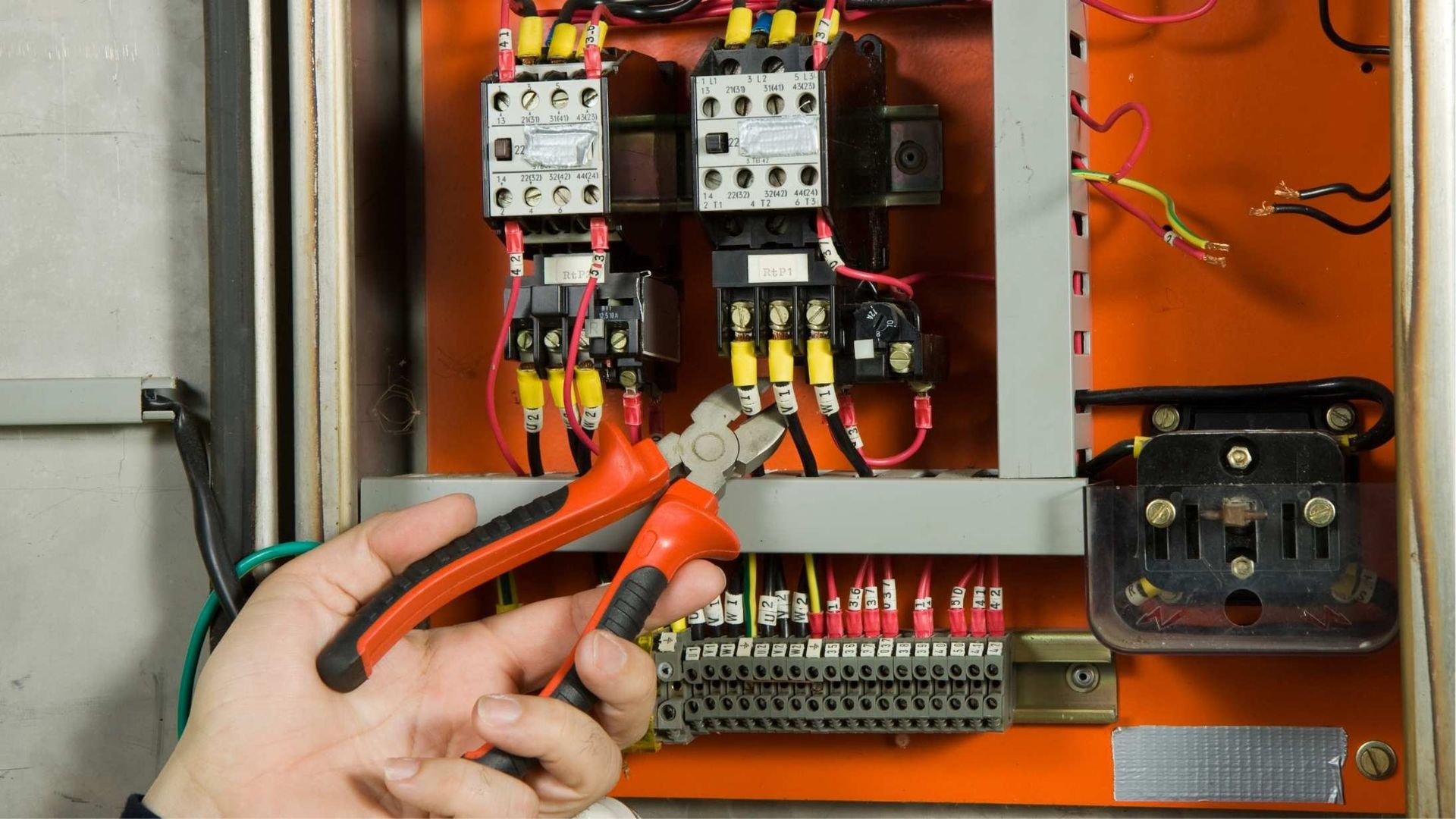 Hands using red pliers to work on electrical wiring inside an orange control panel.
