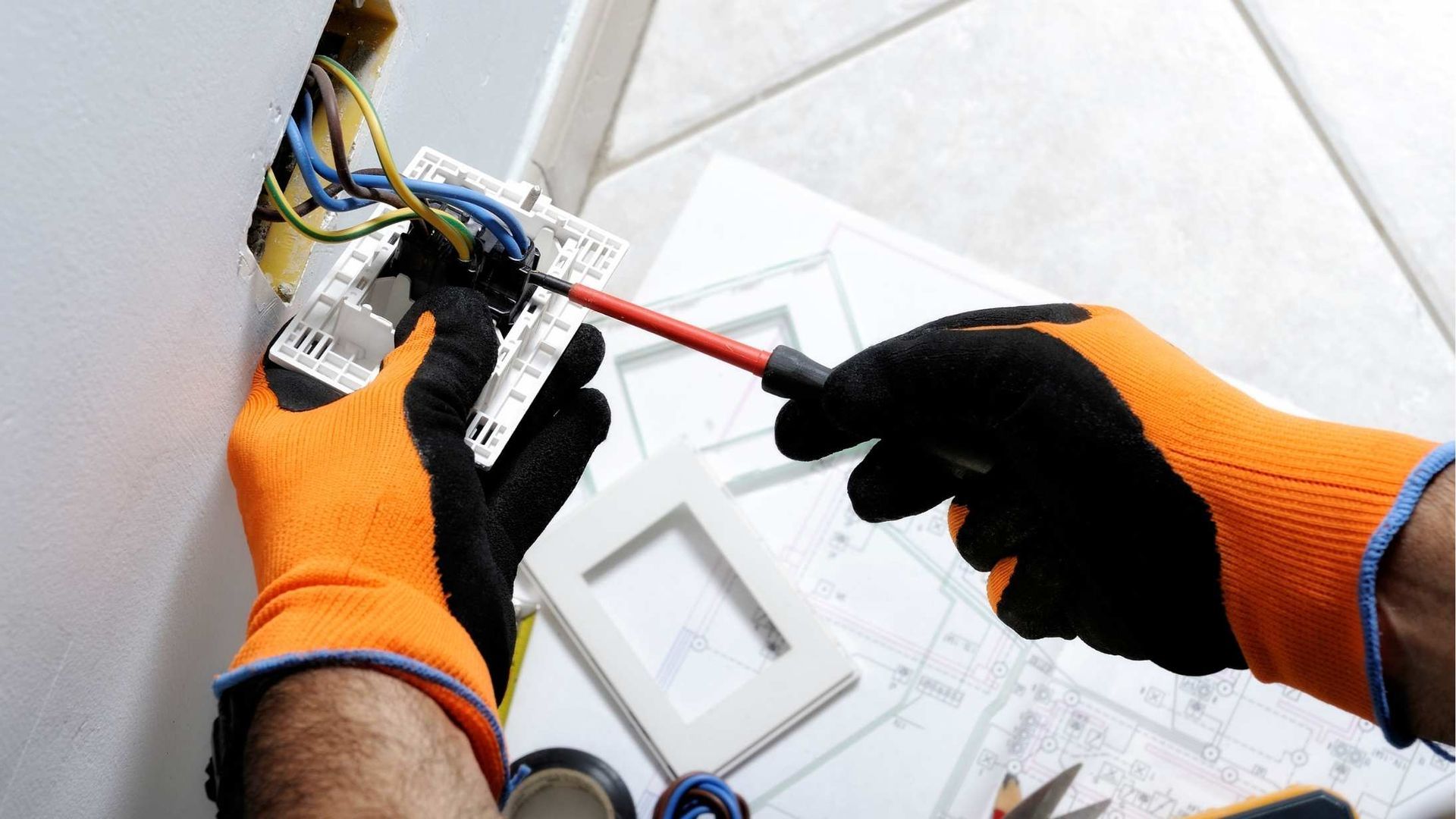 Gloved hands wiring an electrical outlet with a screwdriver on a tiled floor