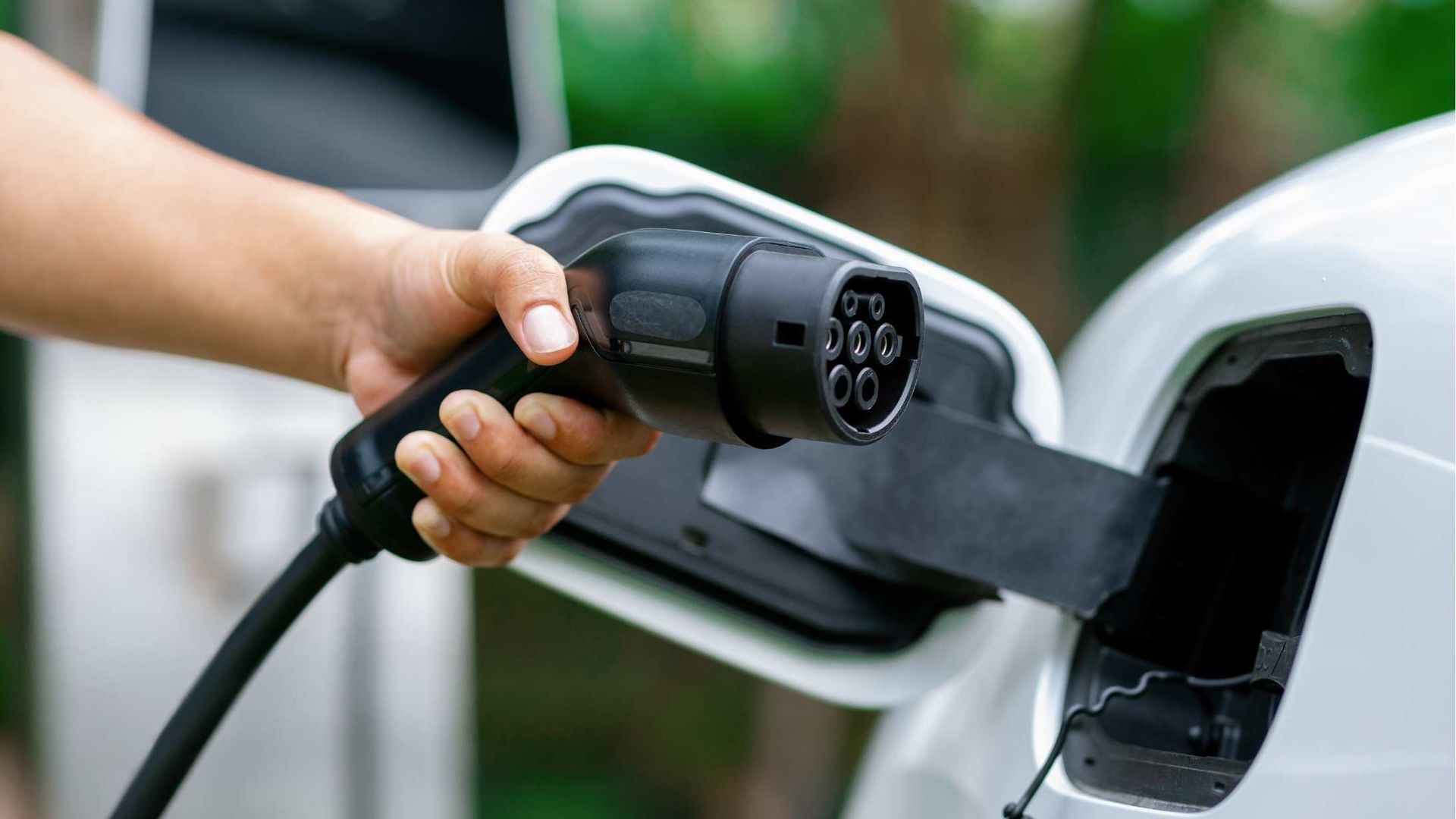 Hand holding EV charging plug connected to a white car at a charging station