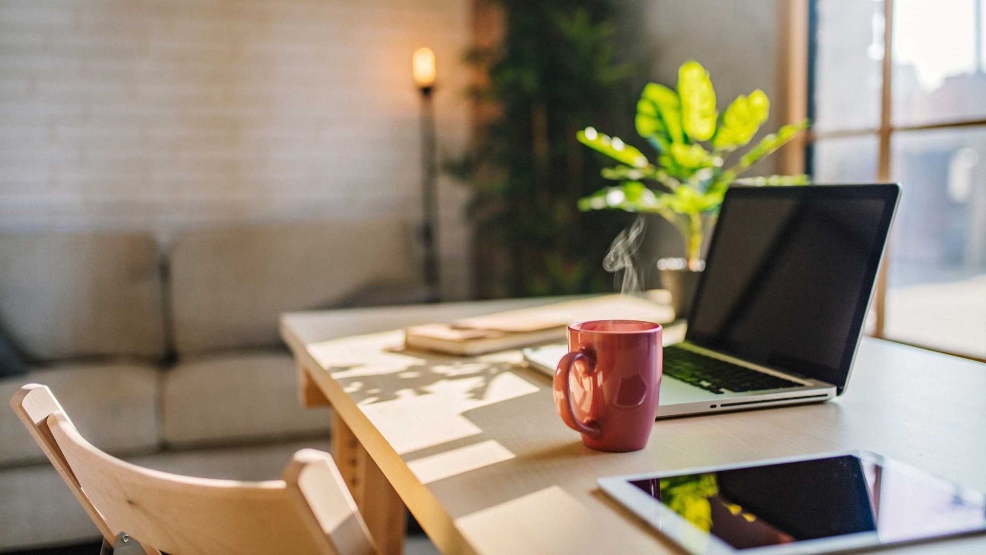 Cozy living room desk with open laptop, red mug, tablet, books, and a potted plant by a sofa and lamp