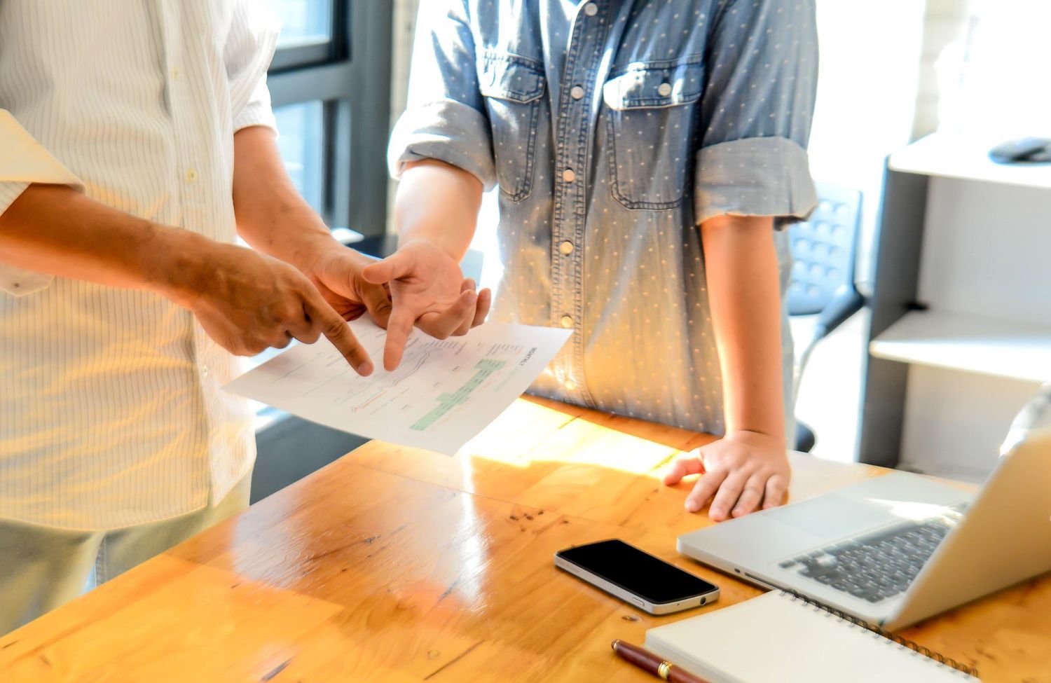 Two people exchanging papers across a wooden table with a laptop and smartphone nearby