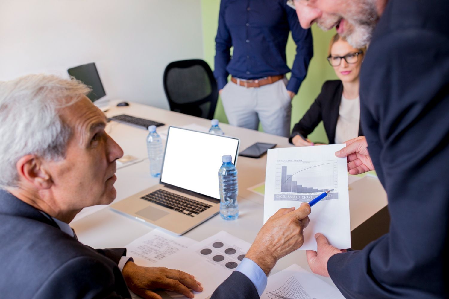 Meeting around a desk with a laptop, papers, and a water bottle, one person pointing at a document.